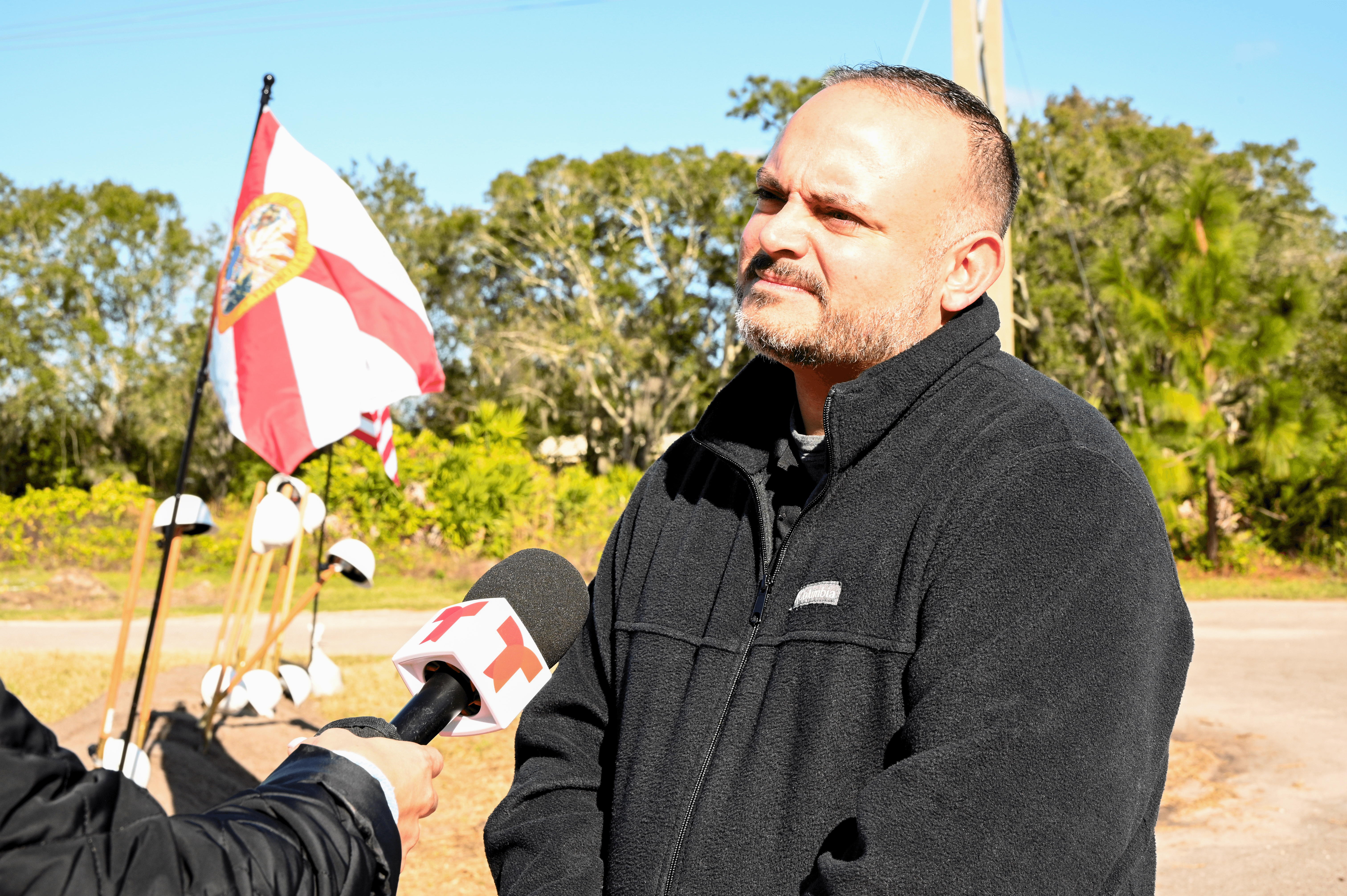 A man answering questions about the groundbreaking. A Florida flag, shovels, and hard hats can be seen in the background